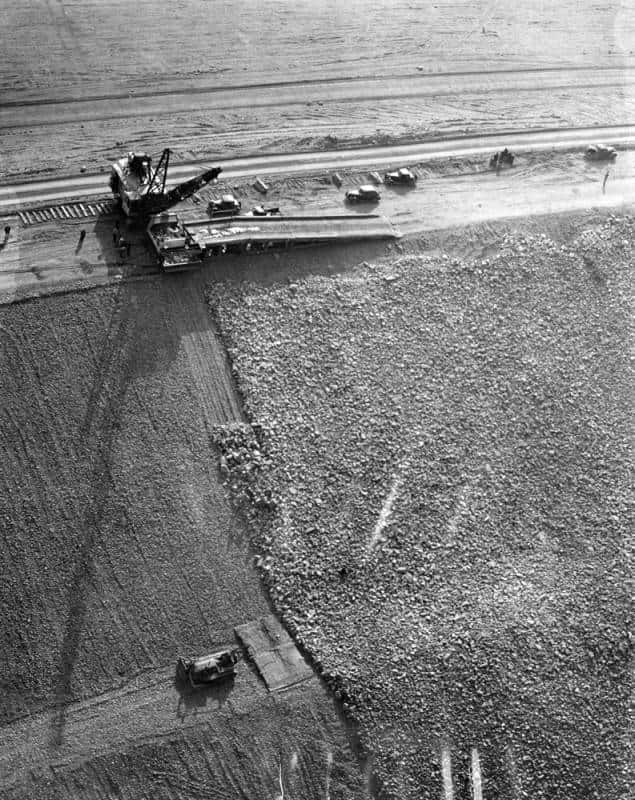 Aerial construction photo from 1940 showing Hansen Dam being built — a derrick and 30-ton truck on a ramp at the top of the frame, with a six-inch gravel layer visible at left and a thick rock layer deposited at right.